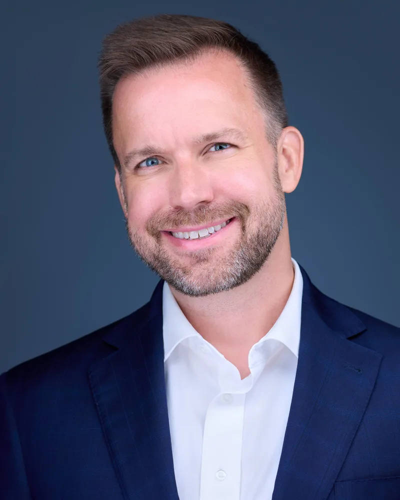 Dan Dzina in a navy jacket and open-collar white shirt, smiling at the camera against a slate-blue studio backdrop