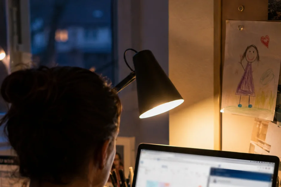 Over-the-shoulder view of a professional at a home office at dusk, calendar on laptop, missed-call notification on phone, hospital portal tab open