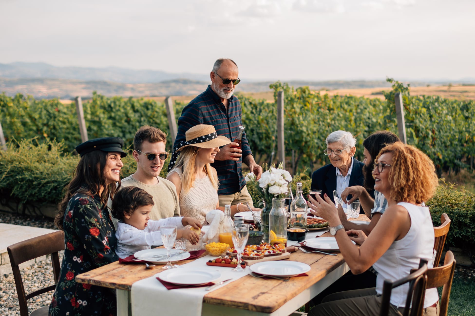 Three generations at a long table in a vineyard, a son serving the family, the grandfather at the end of the table